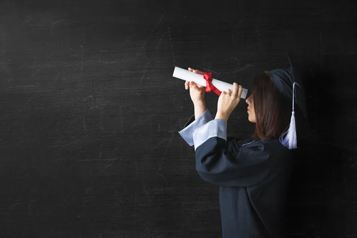 Graduate stands in front of a chalkboard looking through her diploma like a monoscope.